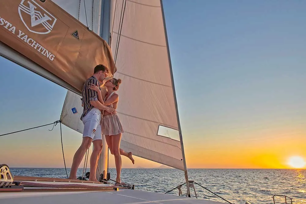 A couple at the bow of a Vista Yachting catamaran, the sail above them and the sun setting over the sea